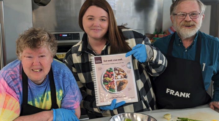 Three people smiling in a kitchen, one holding a Canada’s Food Guide booklet, promoting healthy eating and cooking together.